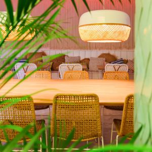 Een sfeervol ingerichte eethoek met natuurlijke verlichting, voorzien van rotan stoelen en een bijpassende bank rondom een houten tafel. De ruimte is versierd met zachte kussens en omgeven door weelderig groen, wat een ontspannen en milieuvriendelijke uitstraling geeft.